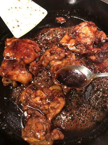 Teriyaki chicken being cooked in a cast iron skillet with sauce turning glossy and thick spoon holding some of the thick sauce and a utensil in the background.