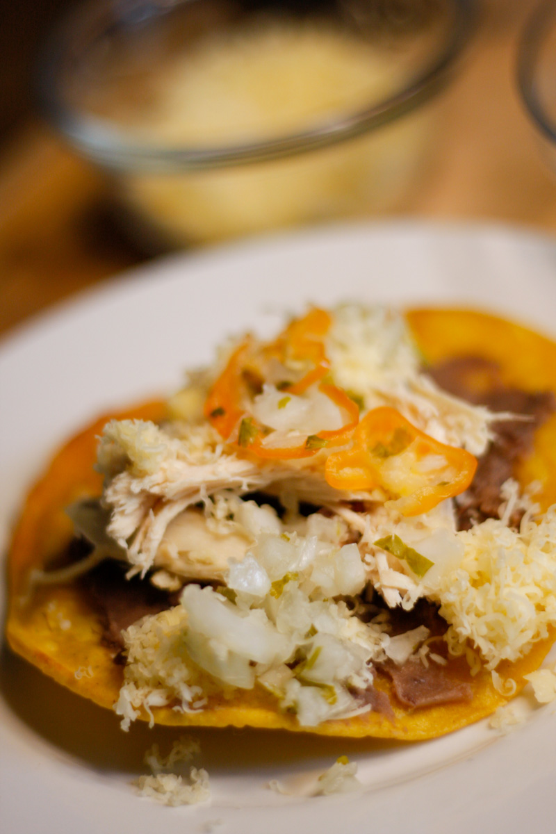 Belizean garnaches which is a corn tostada topped with refried beans, chicken, shredded cheese and relish on a white plate with cheese in the background