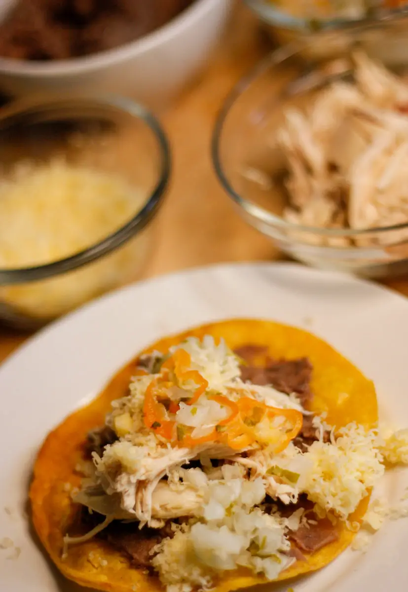 Belizean garnaches which is a corn tostada topped with refried beans, chicken, shredded cheese and relish on a white plate with cheese and chicken in the background