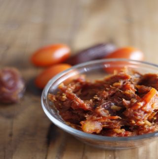 Tomato and date chutney in a glass bowl with tomatoes and dates in the background