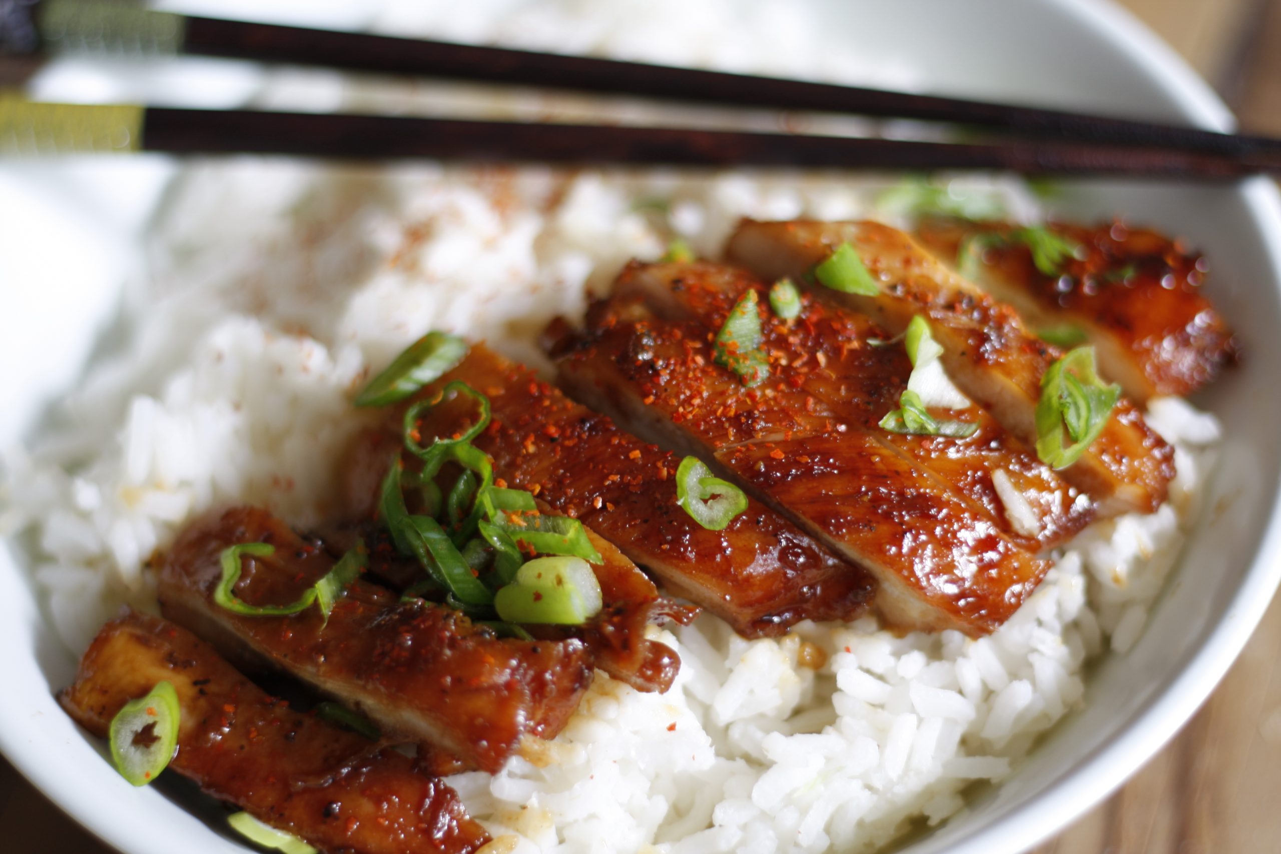 Teriyaki Chicken served with rice and garnished with green onions in a white bowl with chopsticks on top.