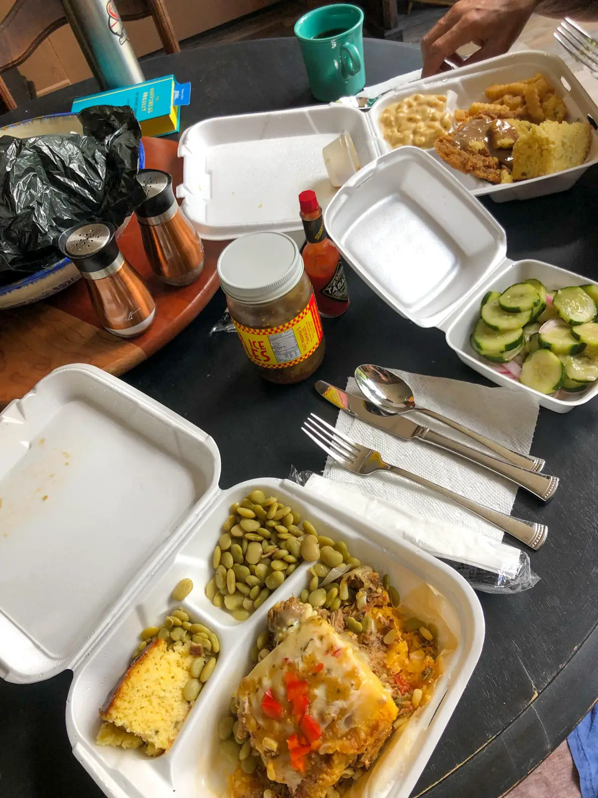 Hot tamale pie, butter beans and corn bread in a styrofoam container, cutlery and napkins, various condiments, cucumber and onions in a styrofoam container and a cup of coffee.