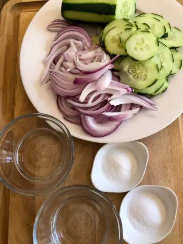 Sliced cucumber and red onion on a white plate, glass bowls filled with water and vinegar, small white dishes with sugar and salt.