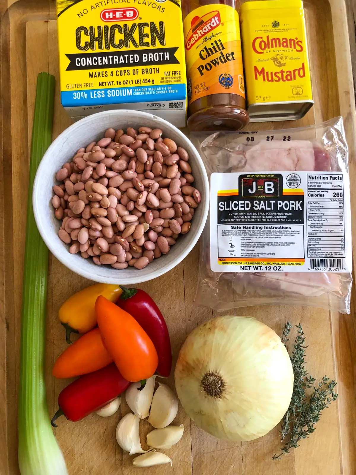 Pink beans in a white bowl, celery stalk, red, orange and yellow sweet peppers, garlic cloves, onion, fresh thyme, package of sliced salt pork, chicken broth, chili powder and Colman's mustard.