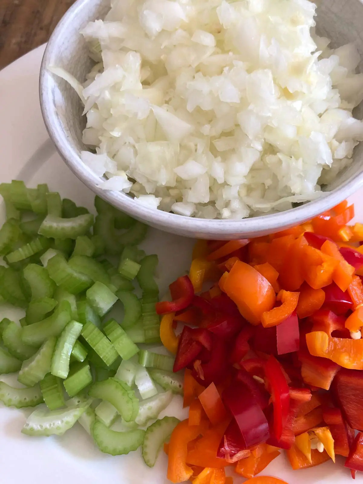 Diced onion in a white bowl, sliced celery and diced sweet bell peppers on a white plate.