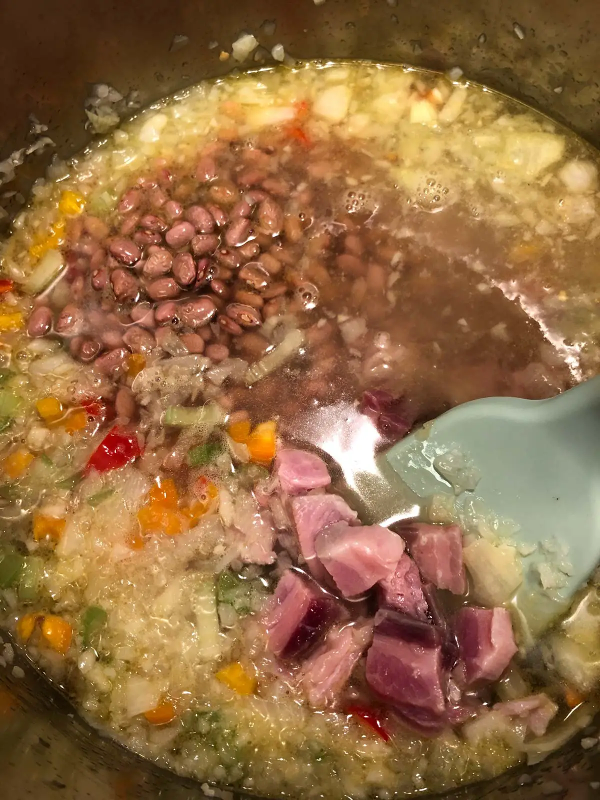 Pink beans, diced and minced vegetables including onions and sweet bell peppers, and diced salt pork in broth in a large pot with a blue utensil.