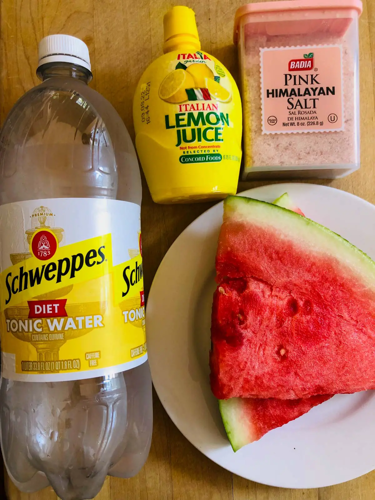 Bottle of Schweppes diet tonic water, lemon juice, pink himalayan salt, and a white plate with watermelon slices.