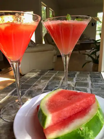 Two martini glasses filled with watermelon mocktail garnished with mint and a white plate with 2 slices of watermelon on a table.