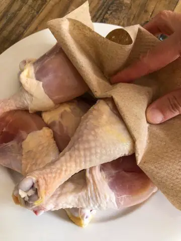 Chicken drumsticks on a white plate with a hand using a brown paper towel to pat dry the chicken.