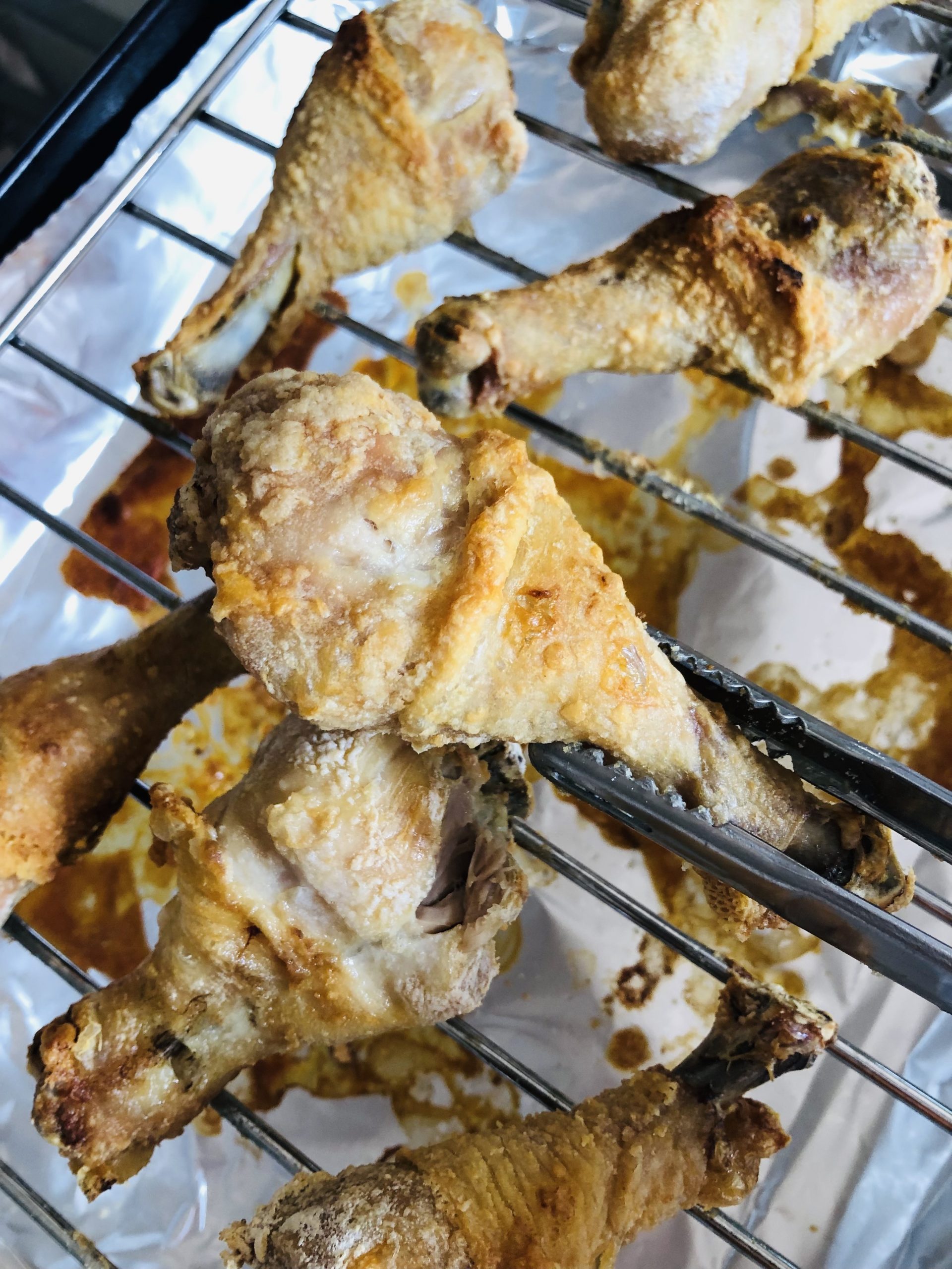 Chicken drumsticks on a wire rack that have been cooked with crispy skin one drumstick being held by tongs closeup.