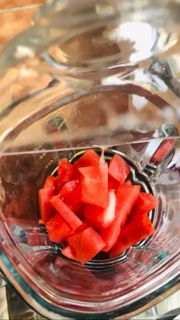 Cubes of watermelon in a blender with a glass dish pouring lemon juice over it.