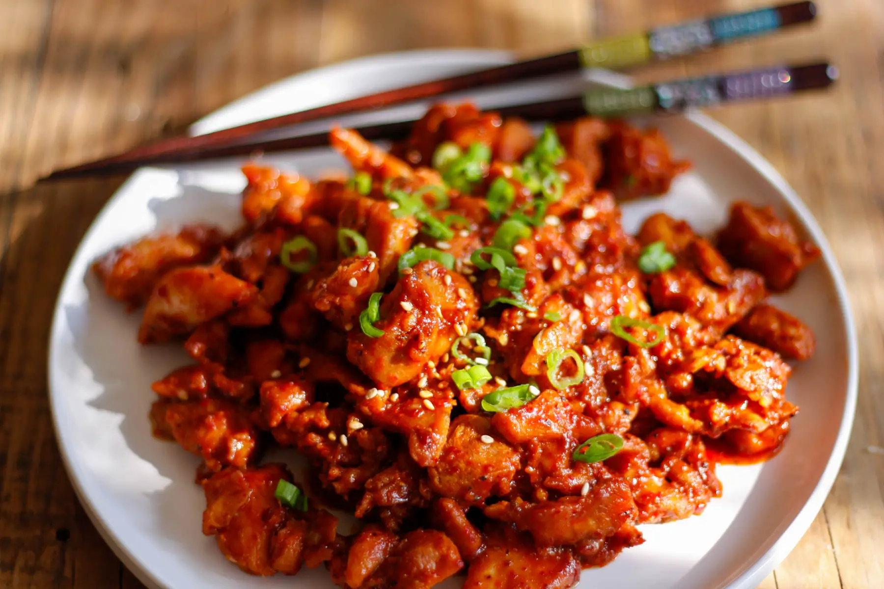 Spicy Chicken Bulgogi garnished with green onions and sesame seeds on a white plate with a pair of chopsticks.