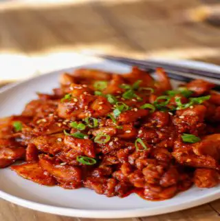 Spicy Chicken Bulgogi garnished with green onions and sesame seeds on a white plate with a pair of chopsticks.