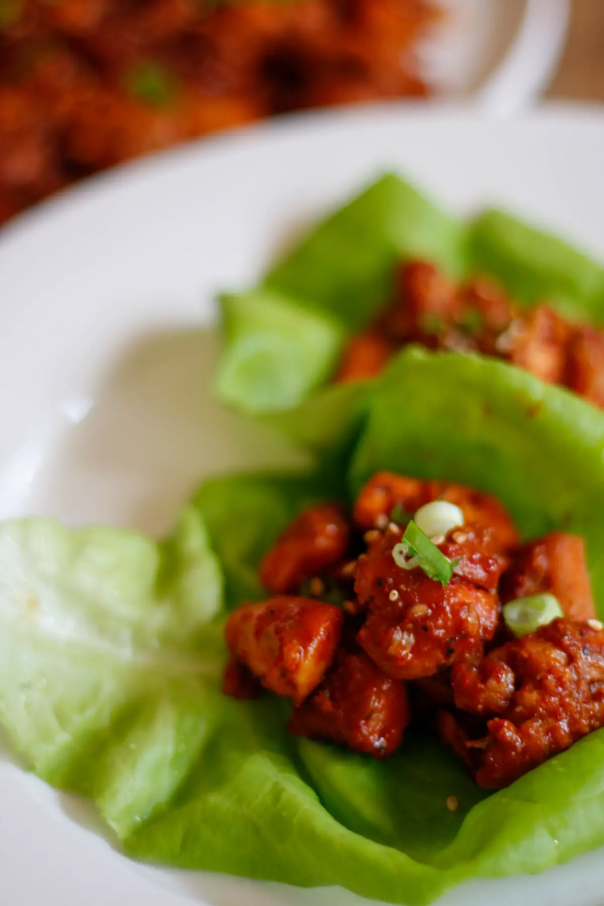 Spicy Chicken bulgogi in lettuce wraps garnished with green onions and sesame seeds on a white plate with more spicy chicken bulgogi in the background.