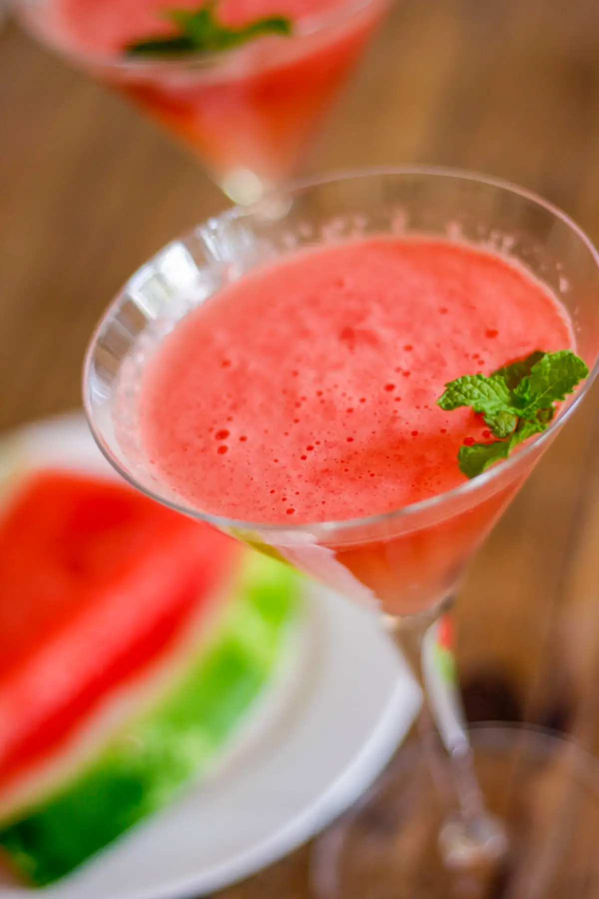 Martini glass filled with watermelon mocktail garnished with mint and one in the background and a white plate with watermelon slices.