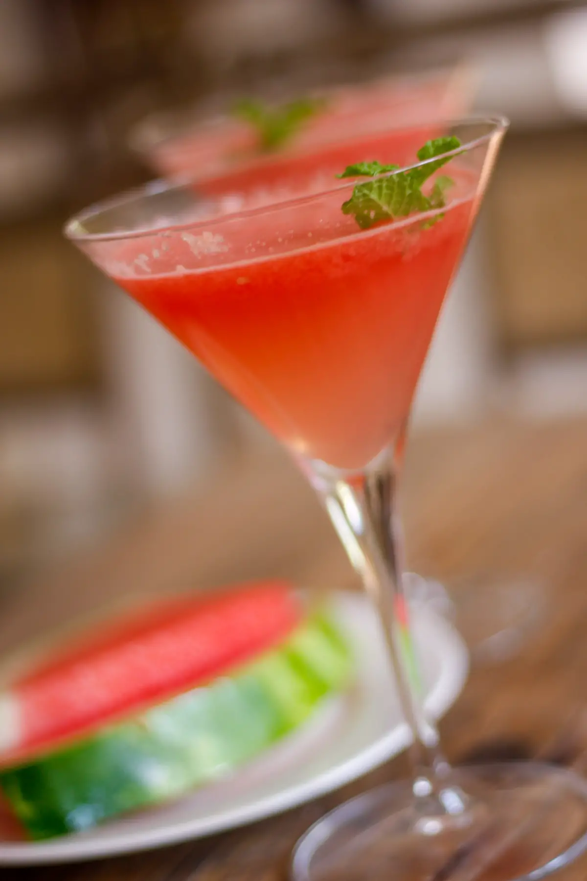 Martini glass filled with watermelon mocktail garnished with mint and one in the background and a white plate with watermelon slices.