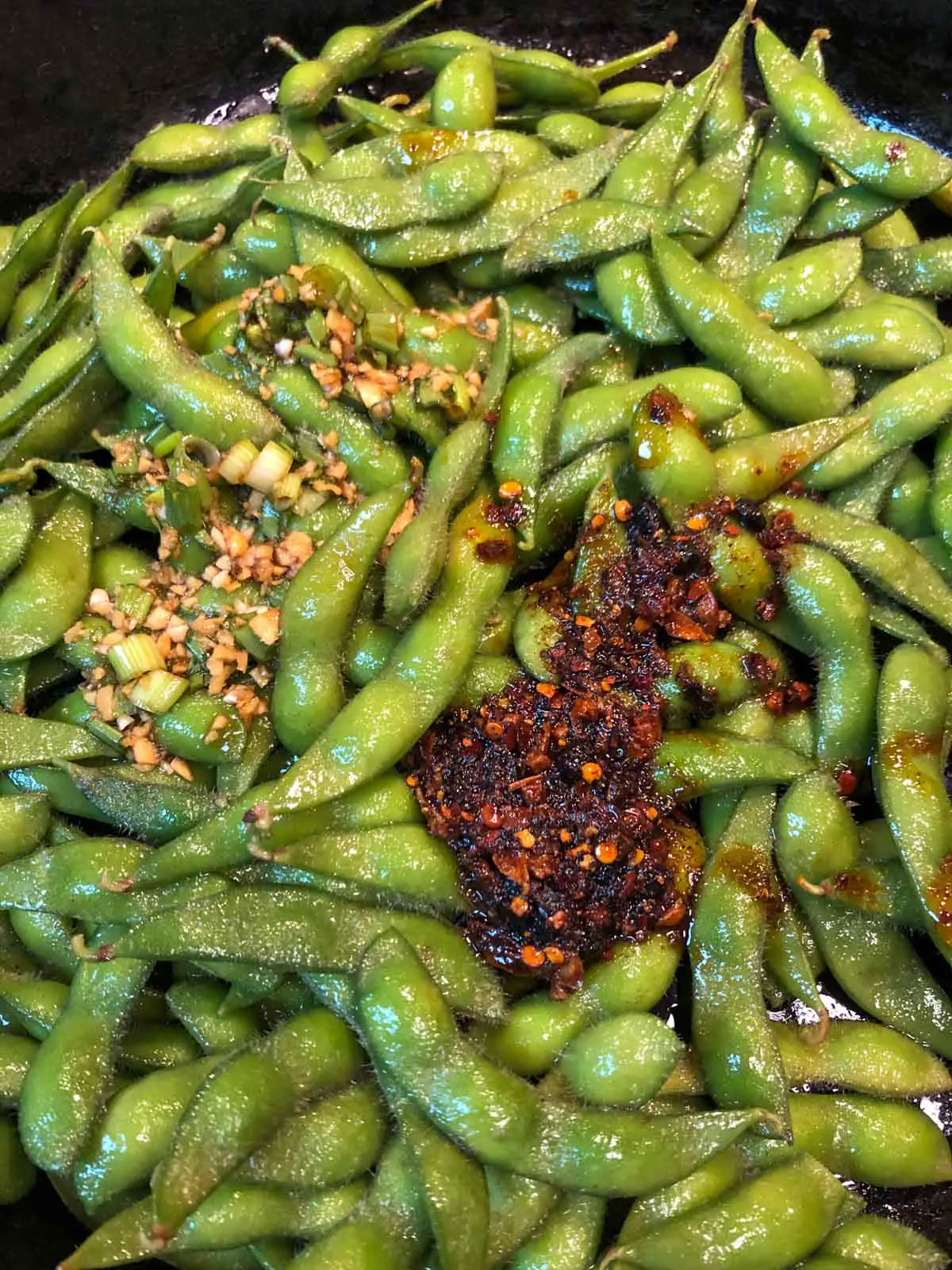 Edamame in their pods with a spicy chili oil on top as well as a ginger sauce with garlic and green onions poured onto the edamame.