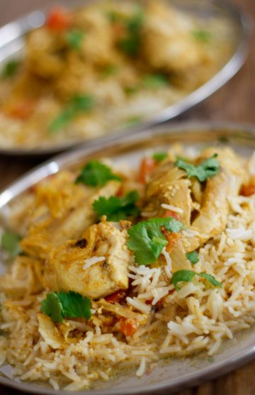 Chicken biryani with chicken pieces, rice, onions, cilantro and tomatoes on a silver plate with a second plate of the same in the background