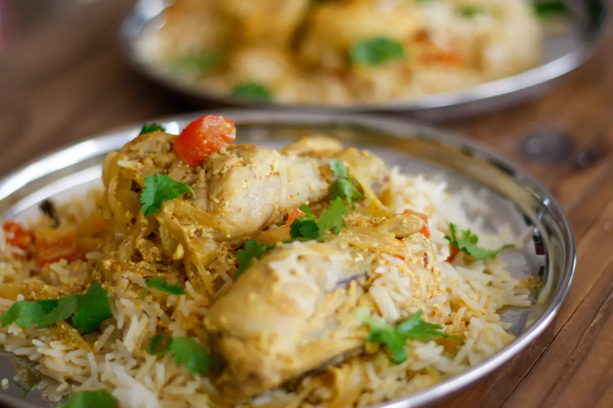 Chicken biryani with chicken pieces, rice, onions, cilantro and tomatoes on a silver plate with a second plate of the same in the background
