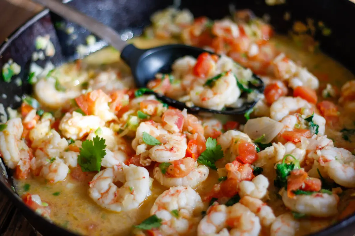 Shrimp with cilantro , diced tomatoes and broth in a cast iron pan with a utensil holding some of the shrimp