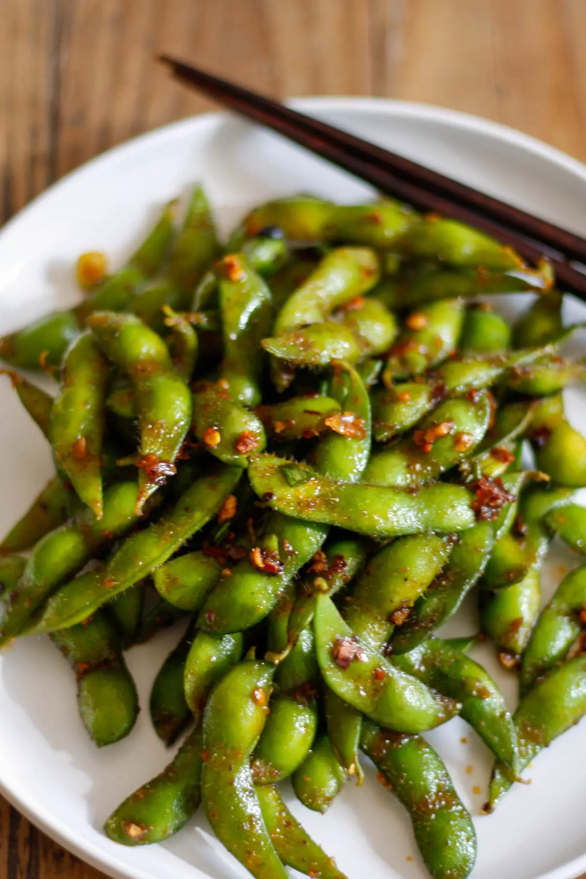 Edamame in the pods with chili sauce on a white plate with a set of chopsticks on the upper right side.