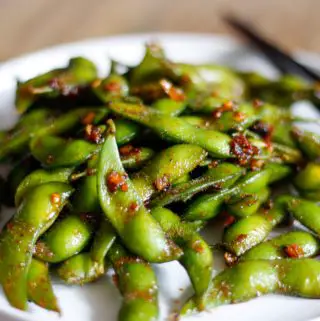 Edamame in the pods with chili sauce on a white plate with a set of chopsticks on the right side.
