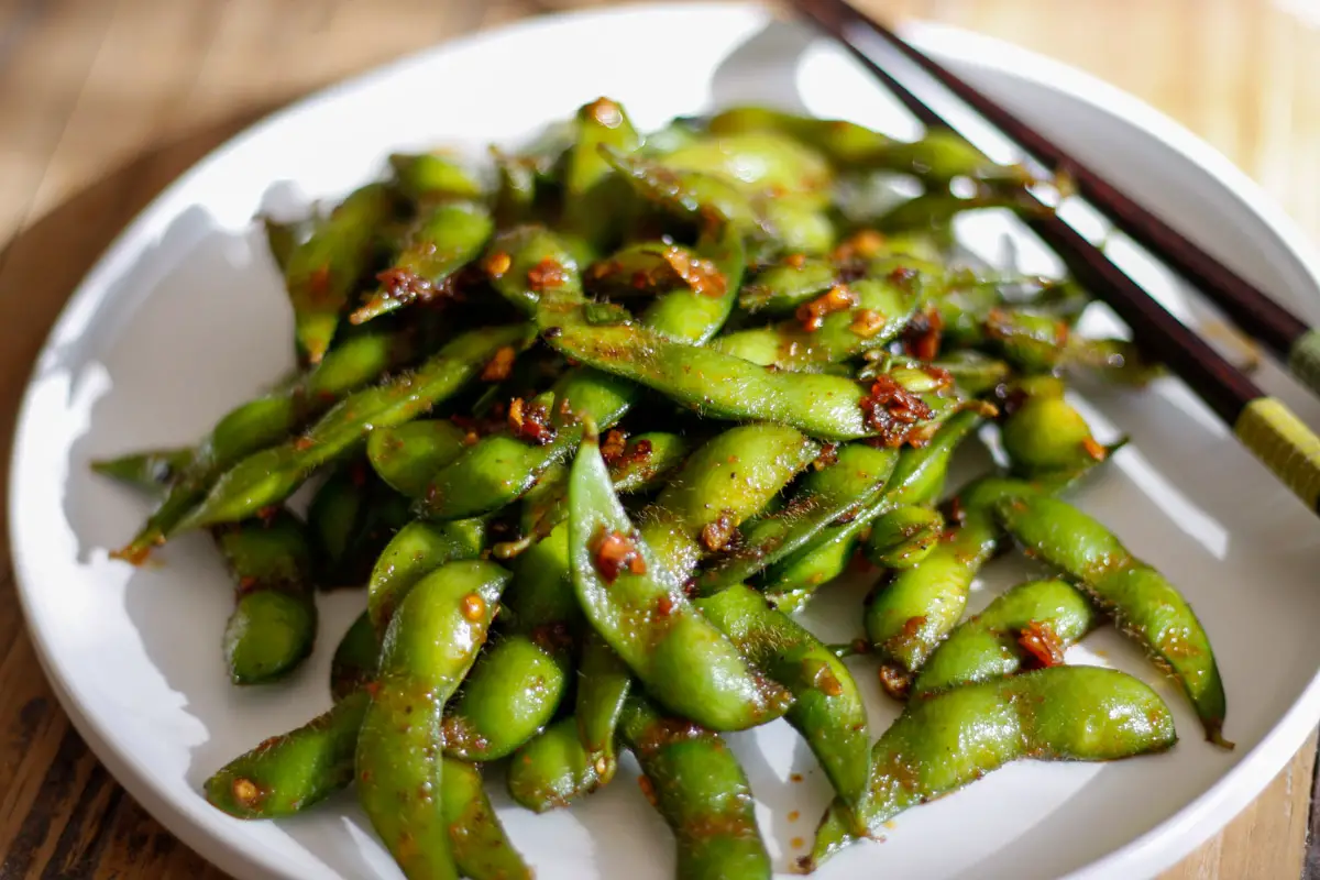 Edamame in the pods with chili sauce on a white plate with a set of chopsticks on the right side.