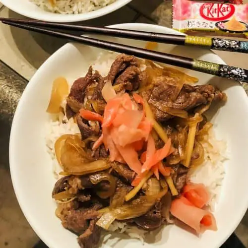 Gyudon which is slices of beef and onion on top of rice and garnished with pickled ginger in a white bowl, chopsticks and Japanese Kit Kat