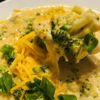 Broccoli and cheddar soup in a white bowl with green onions and shredded cheddar cheese as garnish.