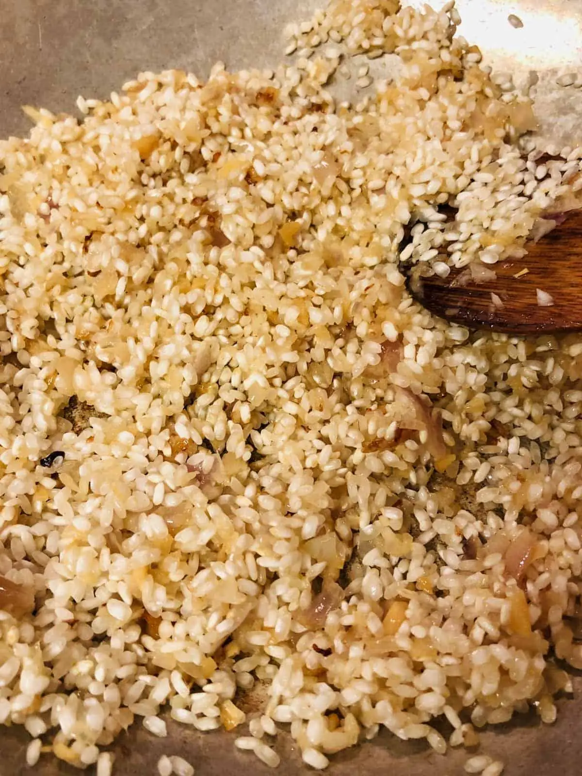 Arborio rice mixed with shallots and garlic in a skillet with a wooden spoon on the right side.