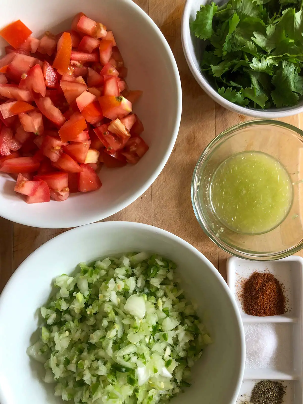 Diced tomatoes in a white bowl, cilantro in a white bowl, lime juice in a glass bowl, minced garlic, chilies, and onion in a white bowl, cayenne, salt, and pepper in a white dish.