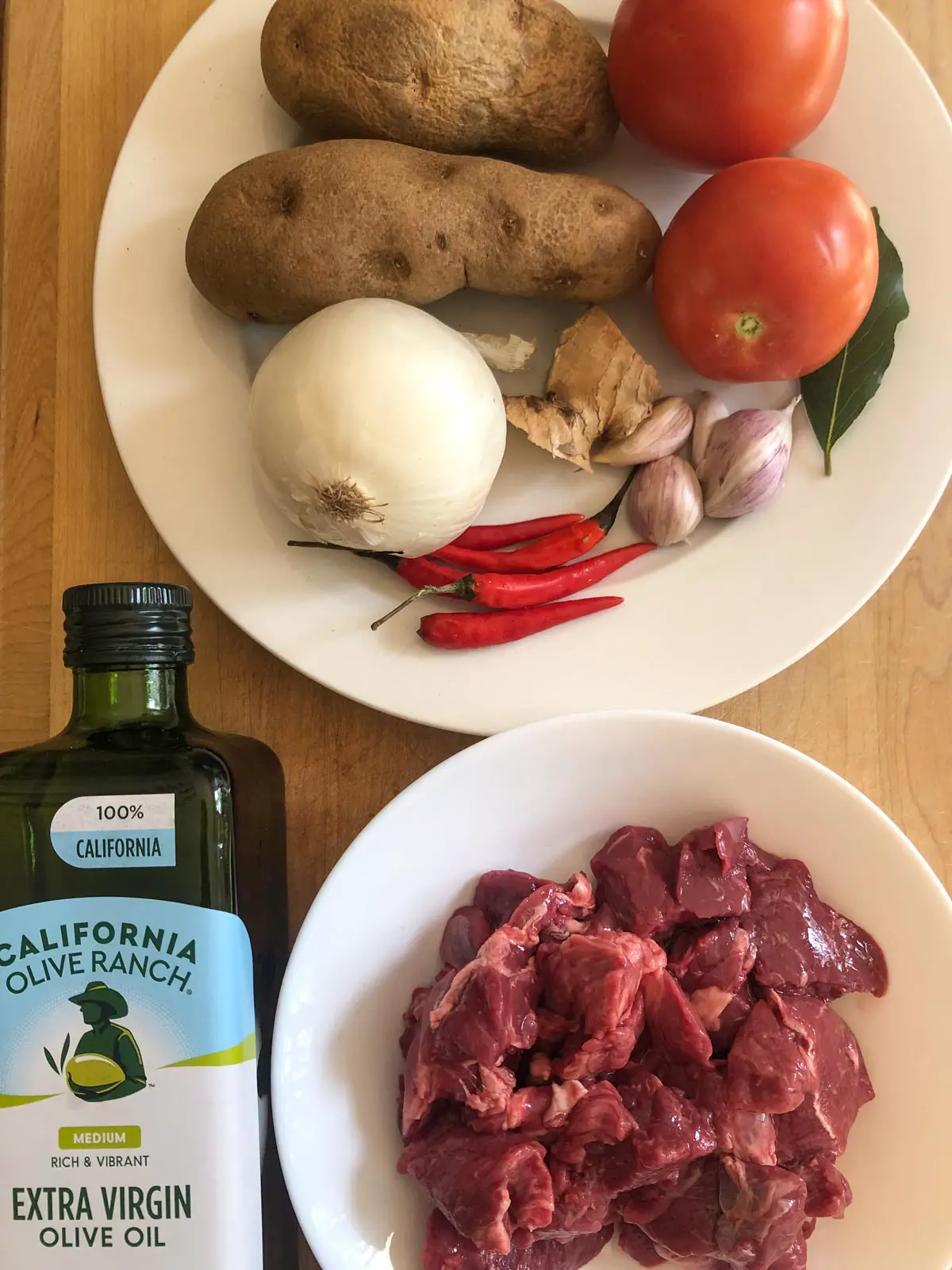 Bottle of extra virgin olive oil, lamb stew meat in a white bowl, and two potatoes, two tomatoes, a white onion, 5 Thai chilies, piece of ginger, 4 garlic cloves and a bay leaf on a white plate.