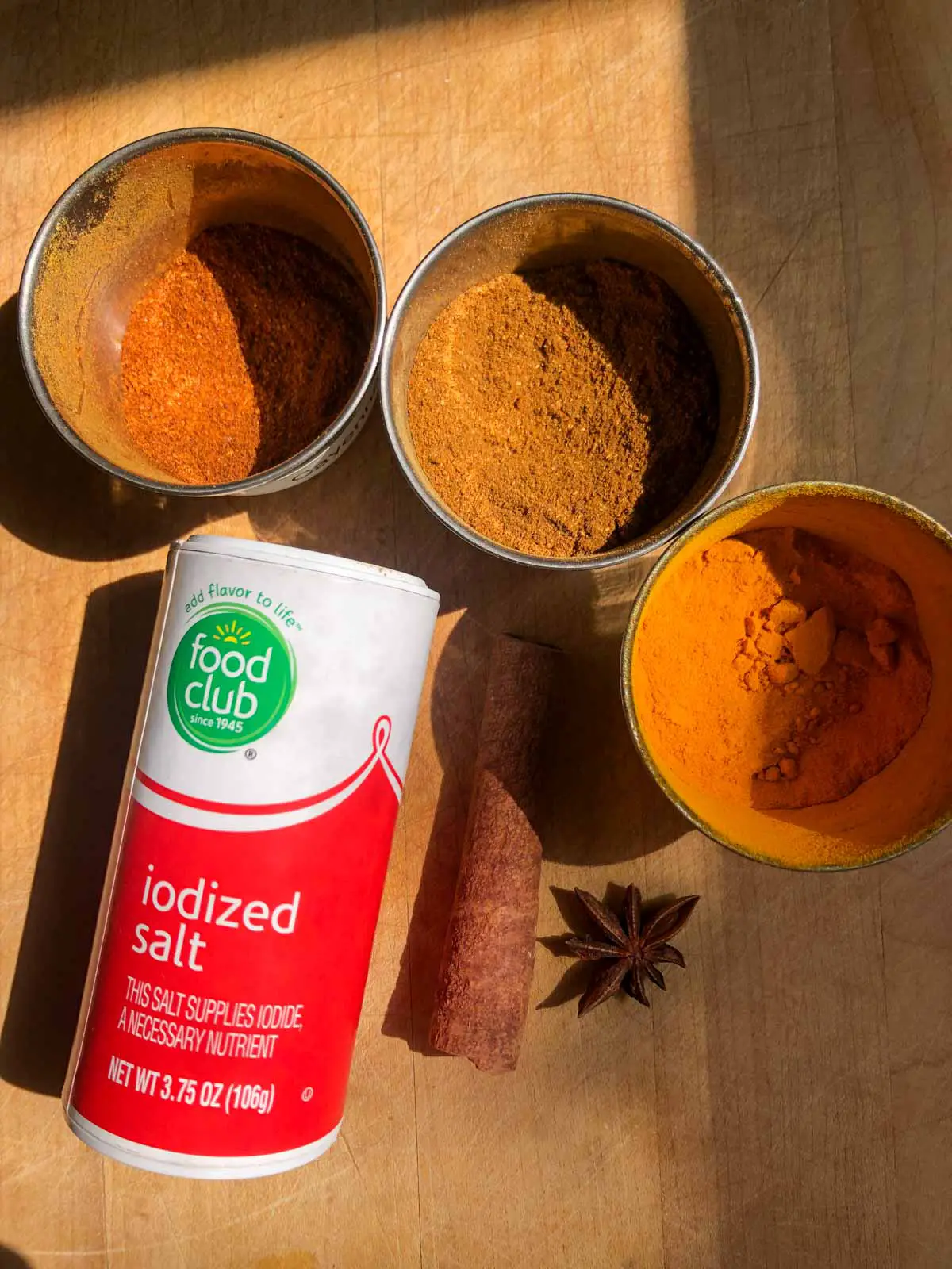 Three metal cups filled with spices one with turmeric, one with garam masala, one with cayenne pepper, a bottle of salt, cinnamon stick and star anise.