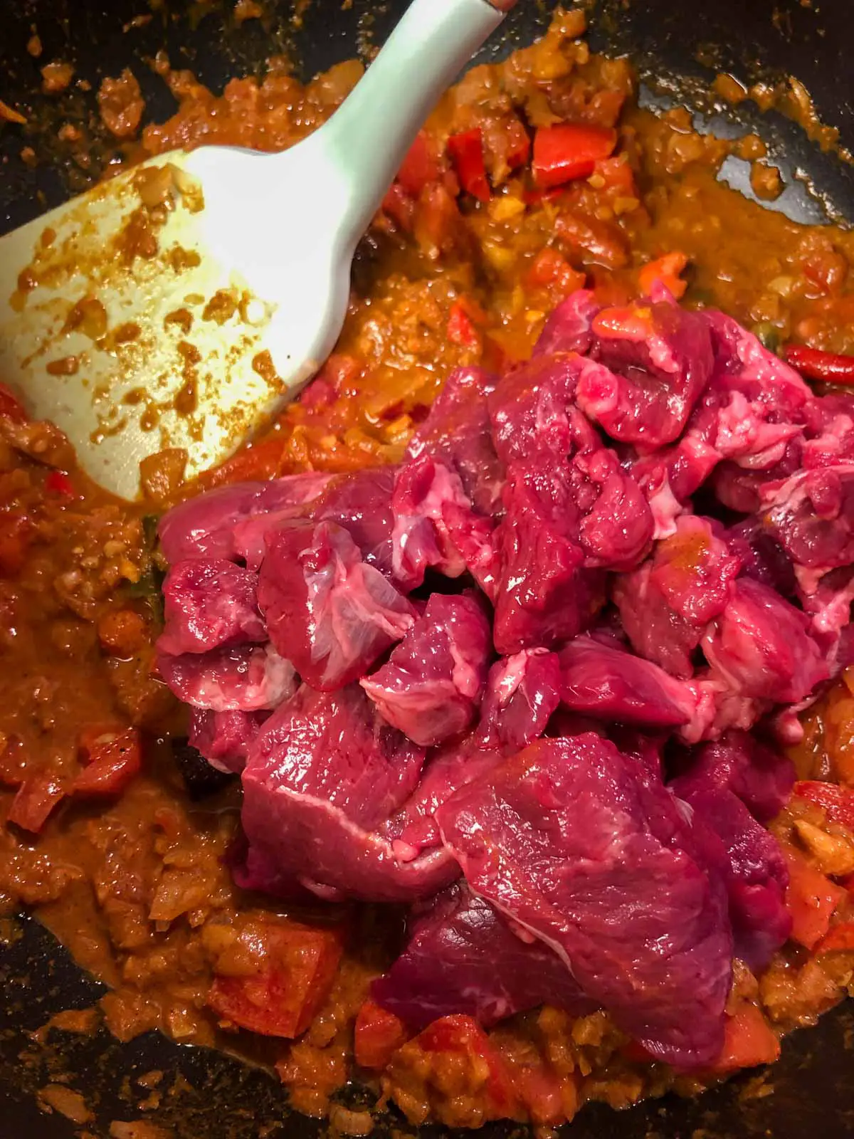 Saucy spice mix with raw lamb for stew meat on top of the spicy sauce with a blue spatula on the upper left hand side.