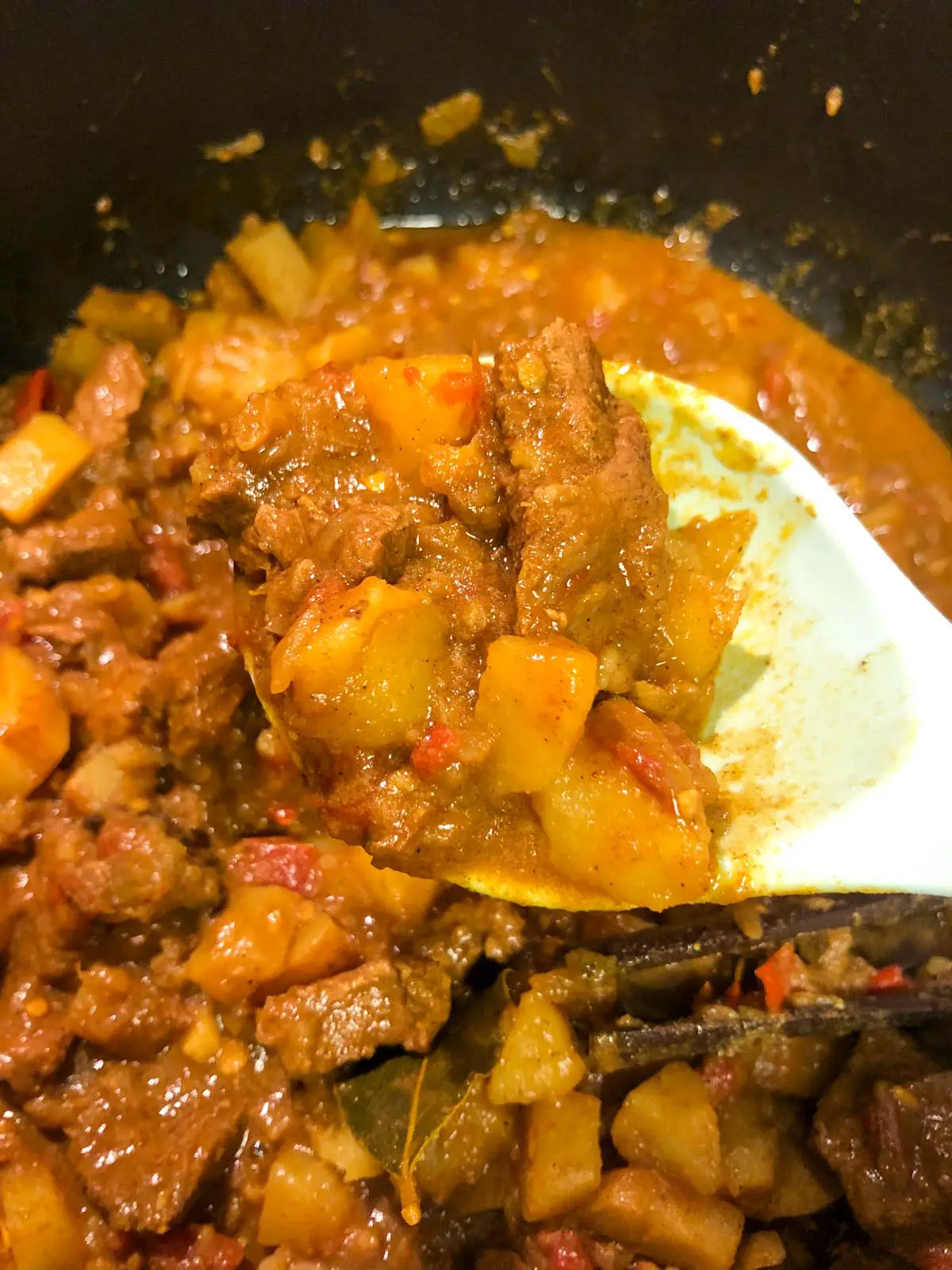 Lamb curry with cut up potatoes, mince chilies, a cinnamon stick and bay leaf, with a spoon containing some of the lamb curry in the foreground.