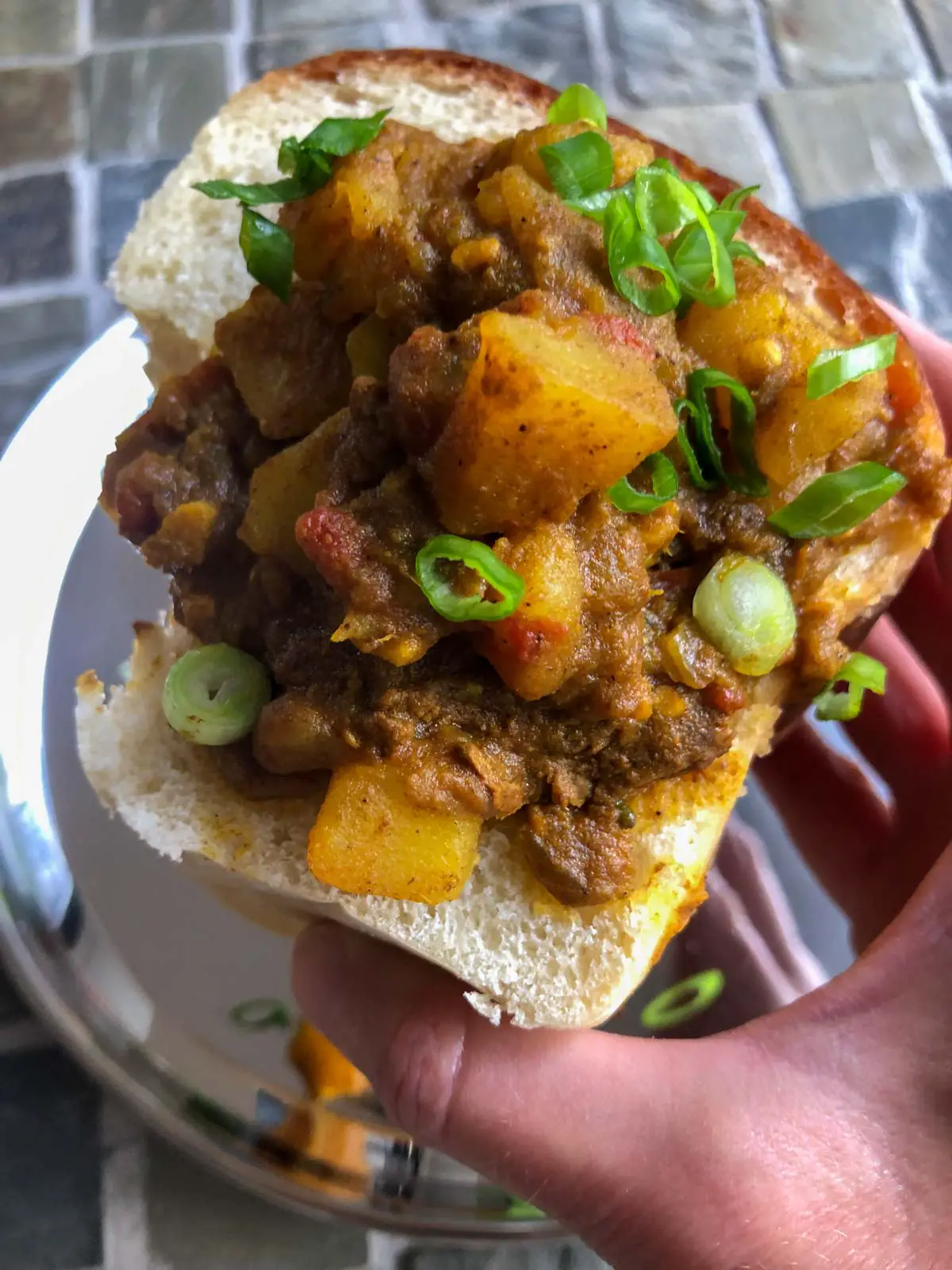 Lamb curry with potatoes and garnished with green onions in a hollowed out piece of white bread loaf being held by a hand.