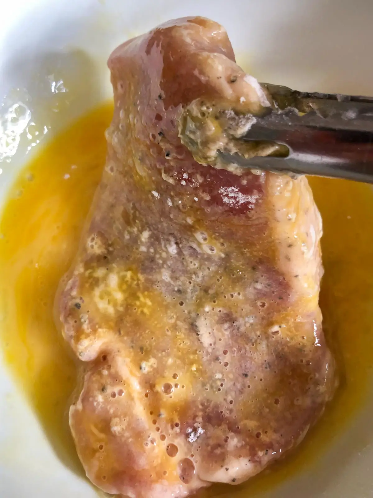 A pork chop coated in flour being dipped into whisked egg that was placed in a white bowl with tongs holding the pork chop.
