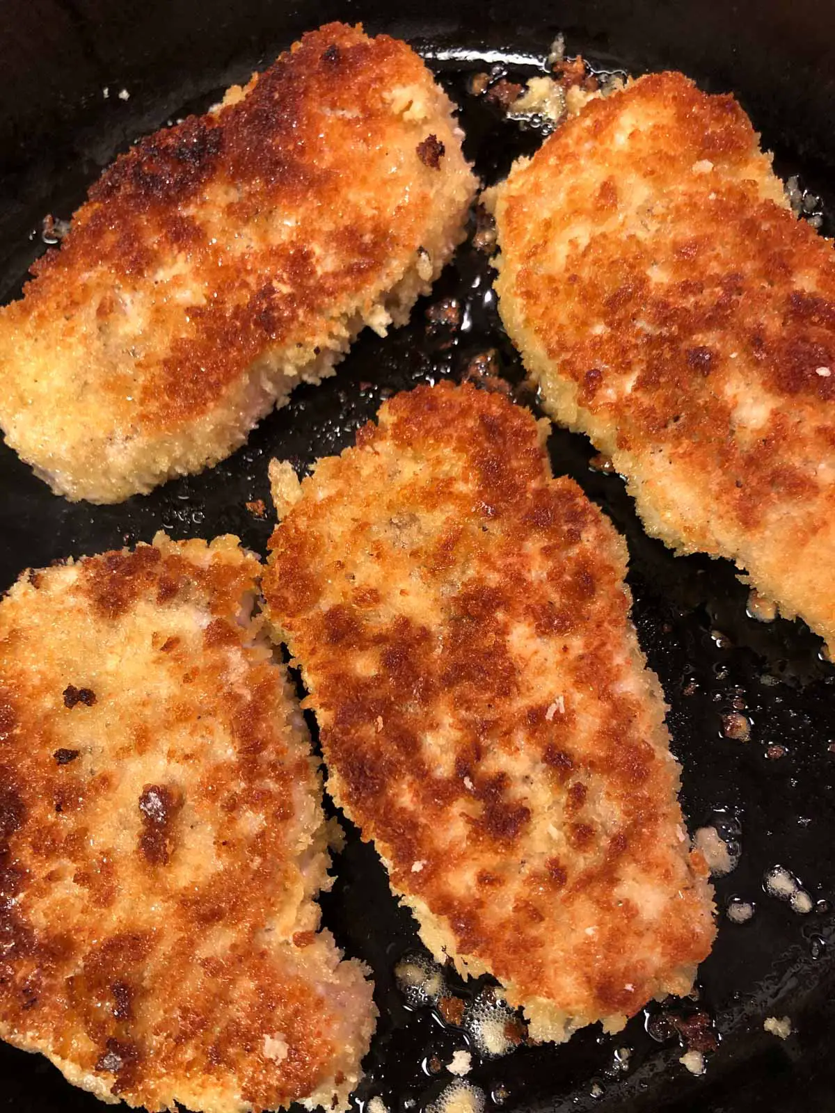 Four brown breaded pork chops frying in oil in a cast iron pan.