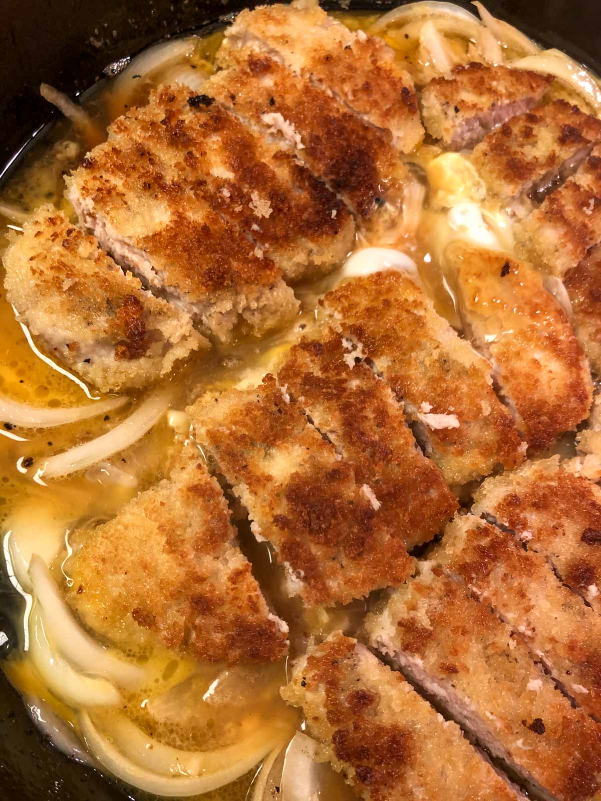 Breaded pieces of pork chops sitting atop onions and surrounded by an egg and broth mixture in a cast iron pan.