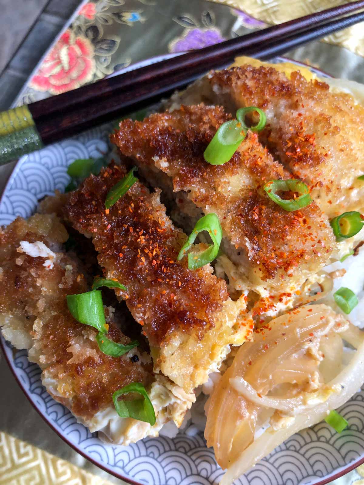 Breaded pork chop pieces on top of rice in a bowl and green onions garnishing the top of the pork chop pieces with a set of chopsticks resting on the left side of the bowl.