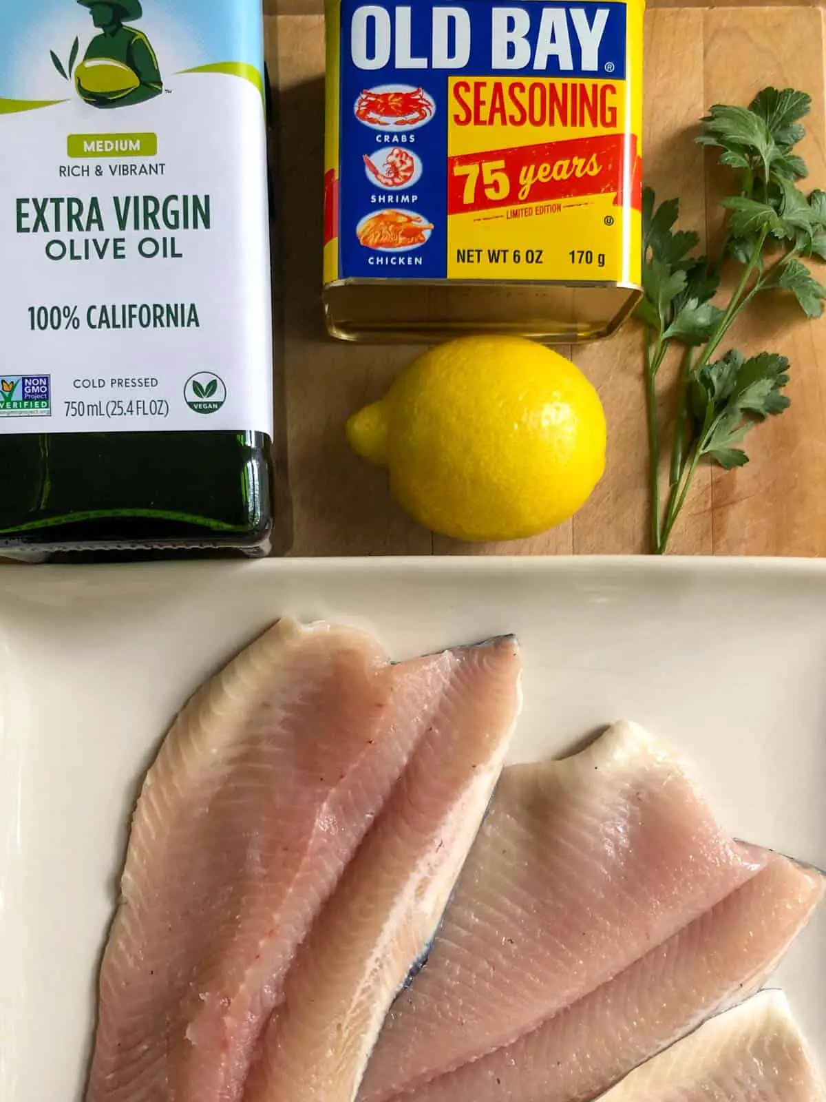 3 filets of rainbow trout on a plate with a bottle of extra virgin olive oil, old bay seasoning, a lemon and Italian parsley above the plate.