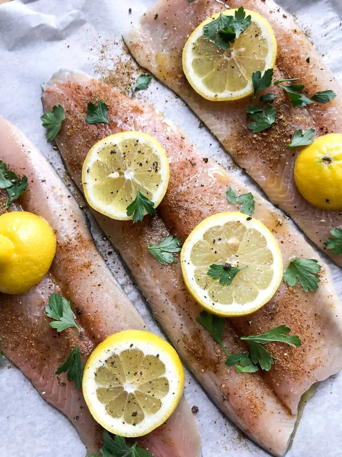 3 filets of Rainbow Trout on parchment paper sprinkled with Old Bay seasoning topped with slices of lemon and Italian parsley.