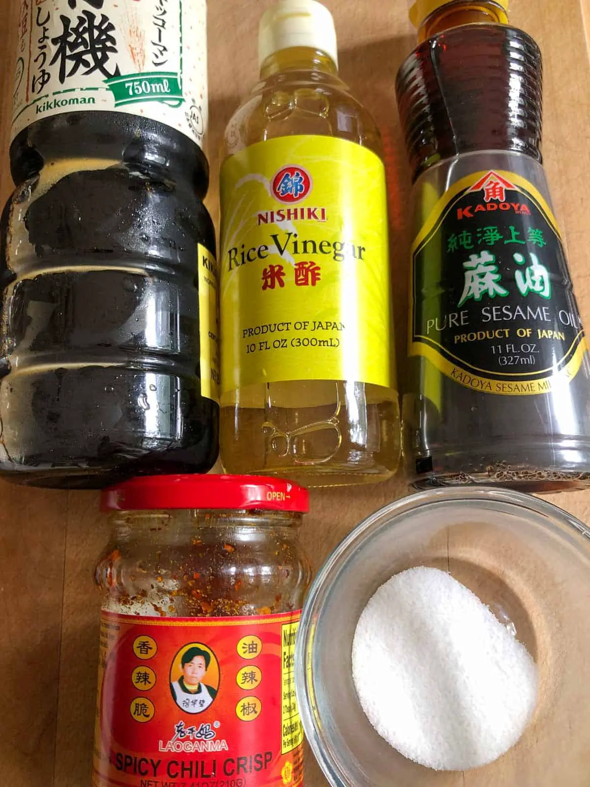 Bottles of soy sauce, rice vinegar, sesame oil, spicy chili crisp, and a glass bowl with sugar