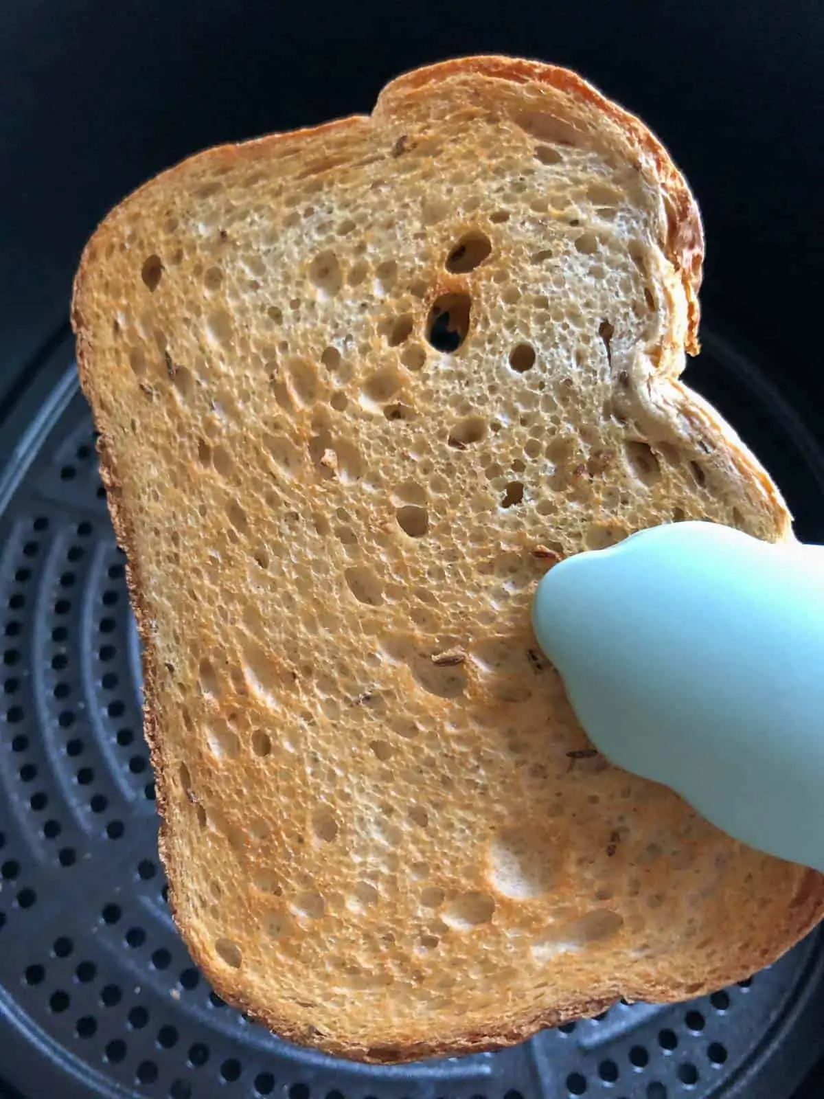Piece of toasted rye bread held by blue tongs with an air fryer basket in the background.