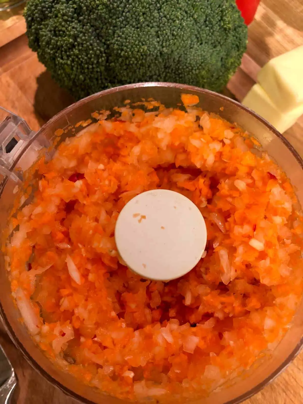 Minced carrot and onion in a food processor with a head of broccoli and some butter in the background.