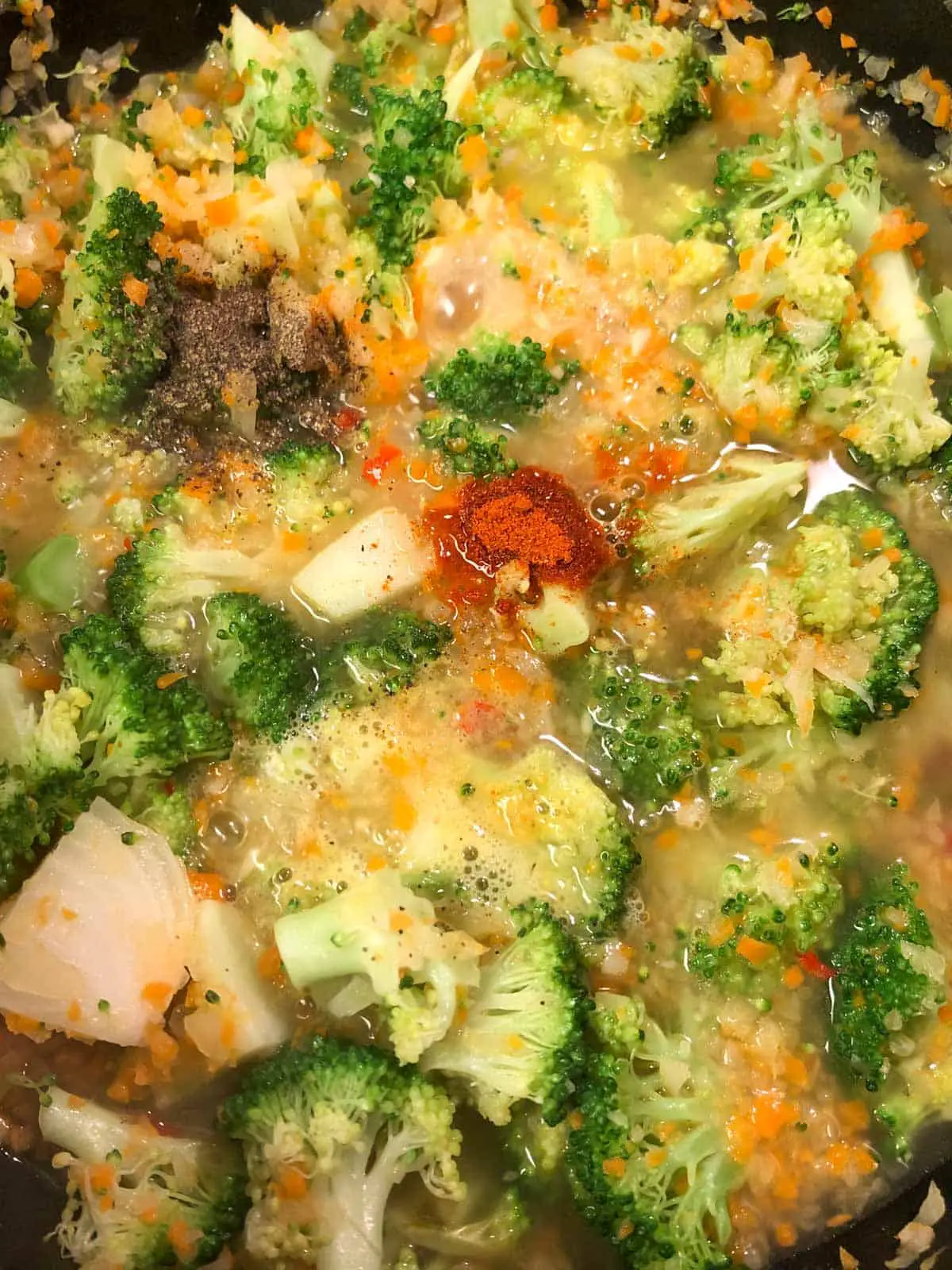 Broccoli florets, minced onion and carrots in a chicken broth, with spices added to the top including pepper and cayenne pepper.