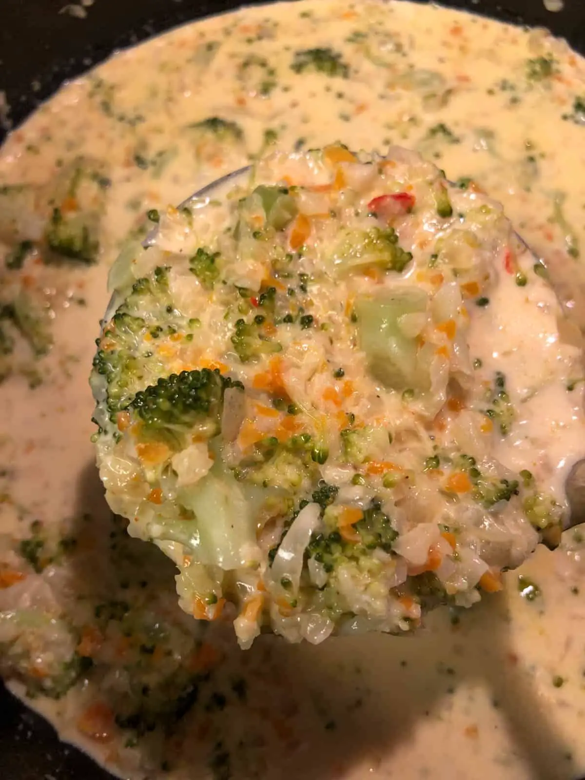 Broccoli and cheddar soup with minced carrots and onion and small pieces or broccoli visible in the soup with a ladle holding some of the soup in the foreground.