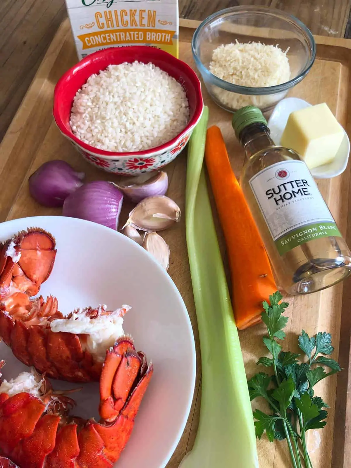 Cooked lobster tails on a white bowl, shallots, garlic, risotto rice in a red bowl, carton of chicken broth, grated Parmesan cheese in a glass bowl, celery stalk, carrot, bottle of Sutter home Sauvignon Blanc, butter in a white dish, and Italian parsley on a wooden cutting board.