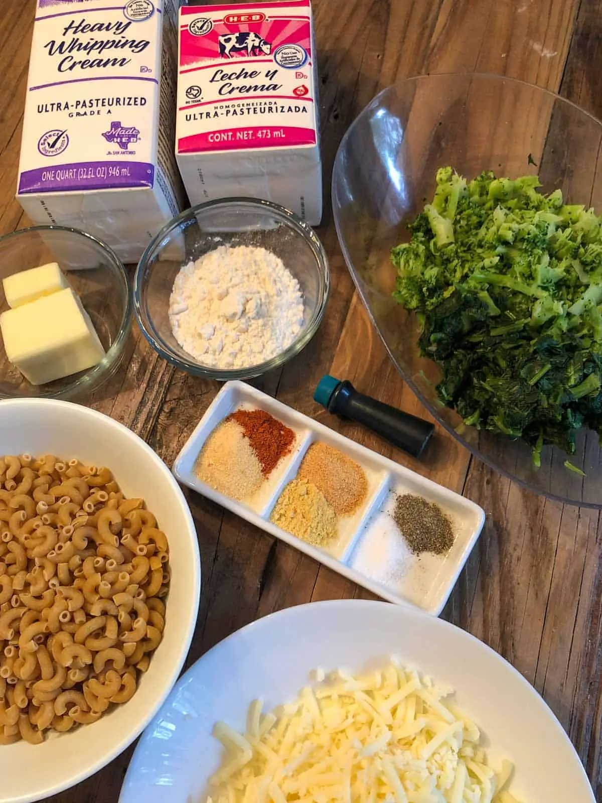 Box of heavy whipping cream, box of ½ and ½, butter in a glass dish, flour in a glass dish, chopped broccoli and chopped spinach in a dish, uncooked macaroni in a bowl, green food coloring vial, white dish with spices including cayenne pepper, onion powder, garlic powder, dry mustard, salt and pepper, white bowl with shredded white cheddar cheese.
