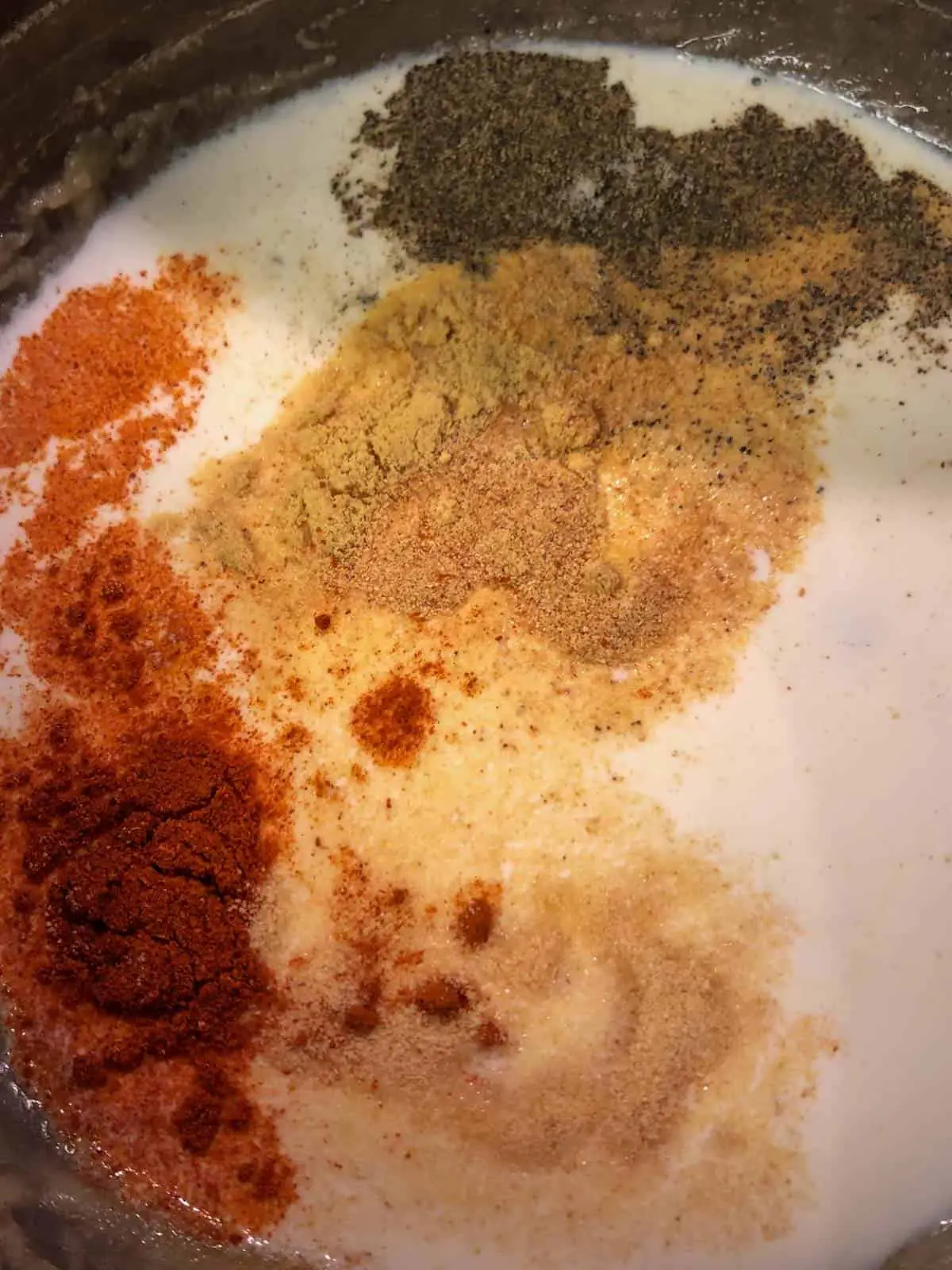 Cream in a saucepan with various spices added to it including salt, pepper, cayenne pepper, onion powder, garlic powder, and dry mustard.